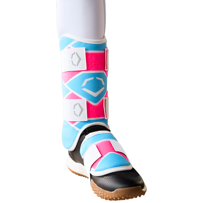 A lower leg wears the EvoShield SRZ-2 Youth Batter's Leg Guard (WB57646) by EvoShield, featuring pink, blue, and white geometric patterns, secured with white Velcro straps and a black and brown sole—resembling youth baseball gear.