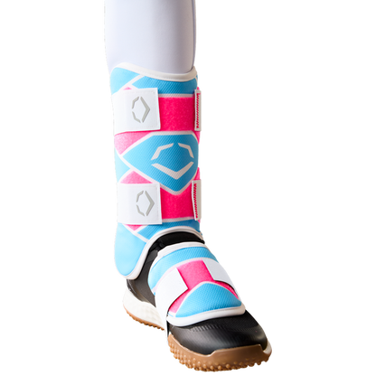 A lower leg wears the EvoShield SRZ-2 Youth Batter's Leg Guard (WB57646) by EvoShield, featuring pink, blue, and white geometric patterns, secured with white Velcro straps and a black and brown sole—resembling youth baseball gear.