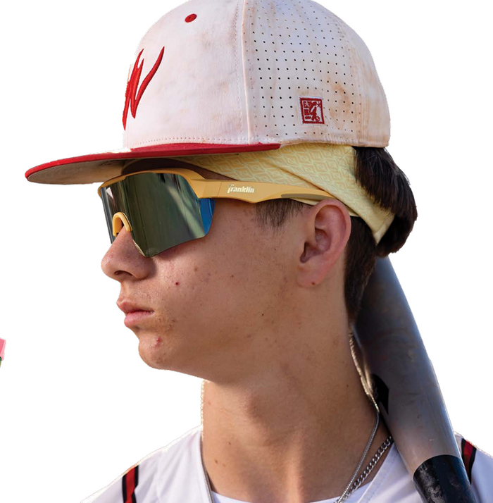 A young baseball player in a white and red cap, wearing Franklin MLB Pro Sunglasses: 24012C, with a yellow headband and a baseball bat on his shoulder, looks to the left.